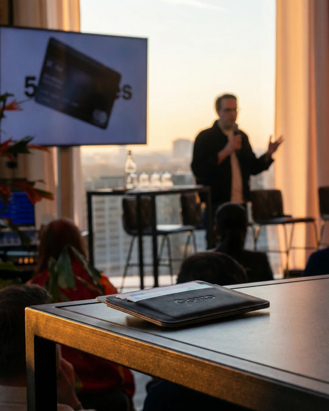 De Qonto-cardholder op een tafel tijdens het event, met een spreker en presentatiescherm op de achtergrond