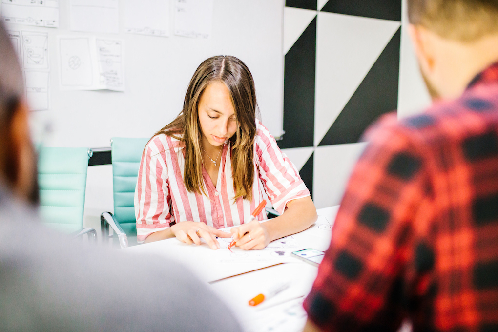 Fiona in a pink and white striped shirt writing with a red pen at a table, with two blurred people in the foreground.