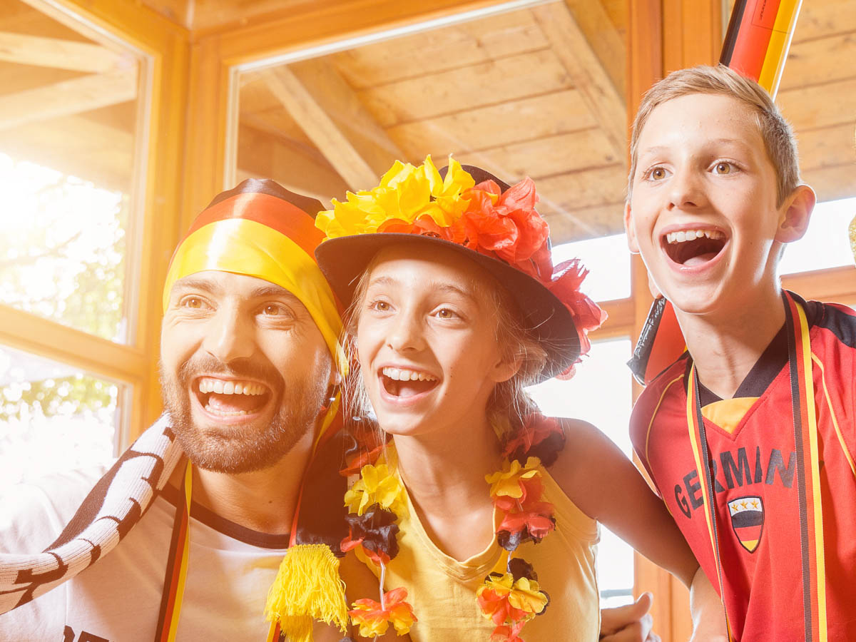 Drei Fußballfans in Deutschland-Fanbekleidung verfolgen begeistert eine Spielübertragung.