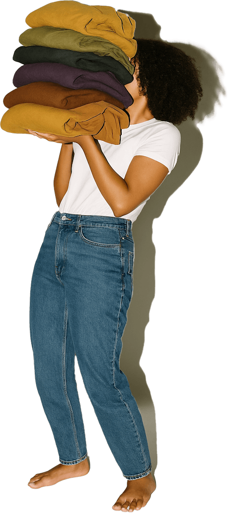 Person in white t-shirt and blue jeans holding a tall stack of folded clothes in earth tones, barefoot.