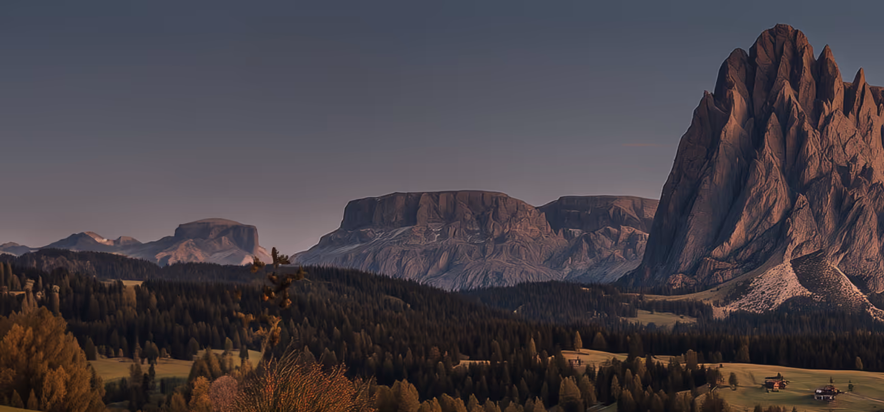 Sunlit rugged mountain peaks towering over dense forest and scattered houses in a valley at dusk.