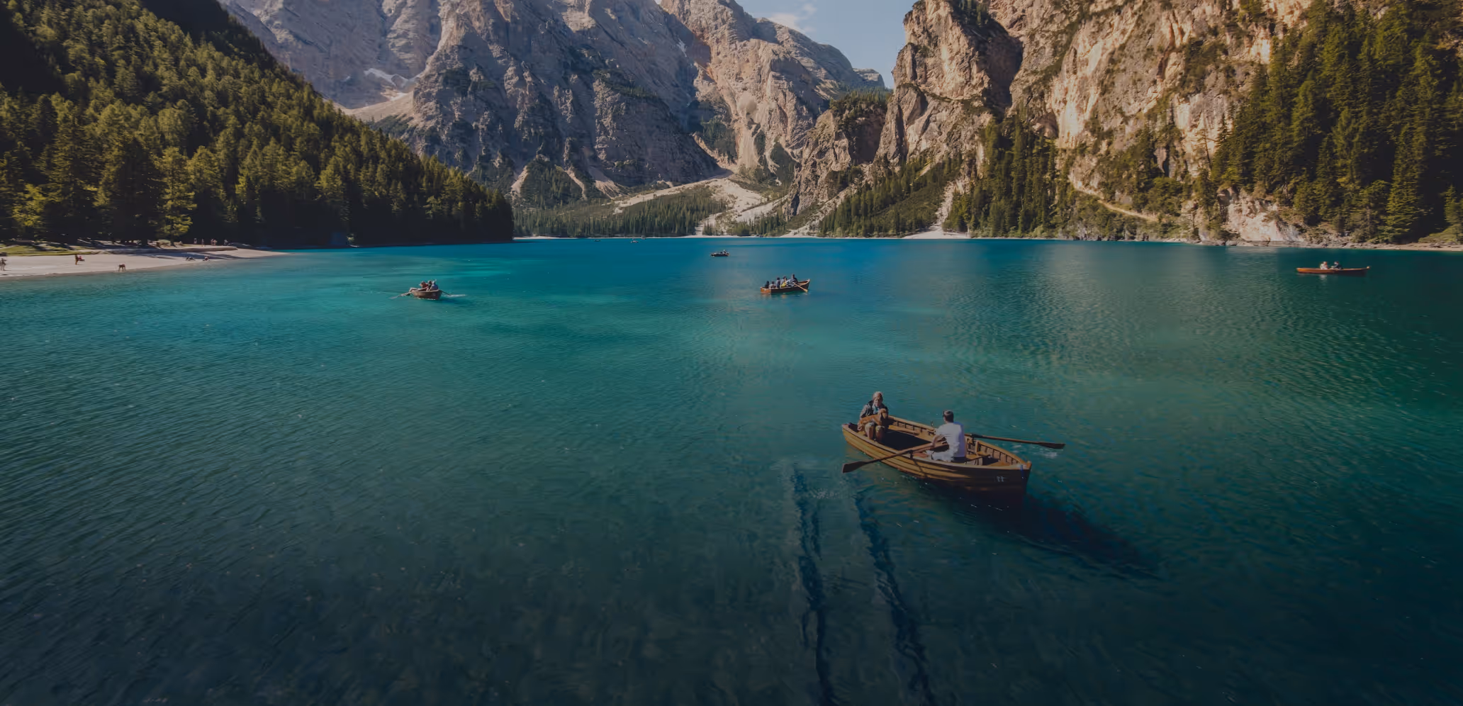 People rowing wooden boats on a clear turquoise mountain lake surrounded by pine forests and rocky cliffs.