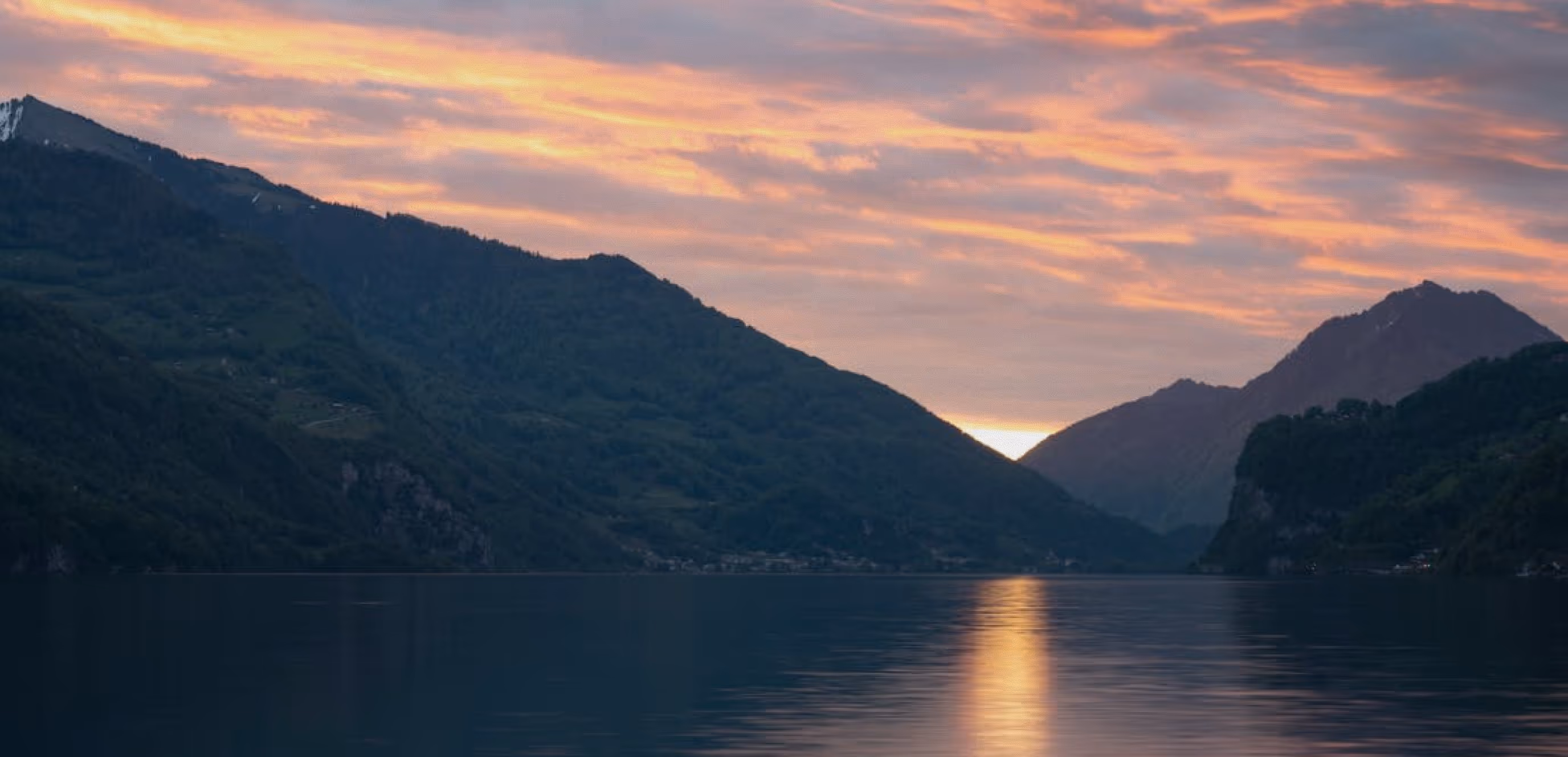 Sunset over a calm lake surrounded by forested mountains, with the sun reflecting on the water.