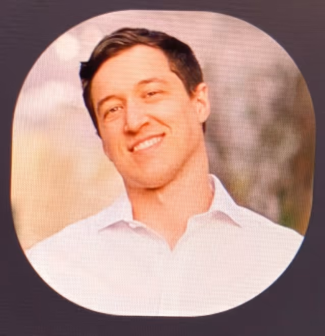 Young man smiling, wearing a white collared shirt, with a blurred outdoor background.