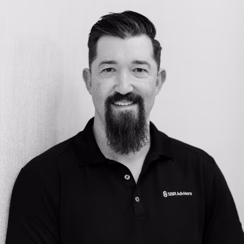 Man with dark hair and a long beard smiling, wearing a black polo shirt with SBIR Advisors logo.