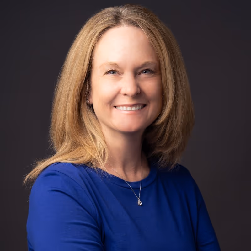 Smiling woman with shoulder-length blonde hair wearing a blue top and a necklace against a dark background.