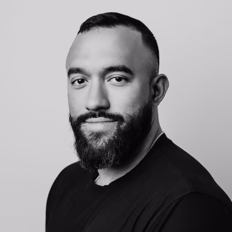 Black and white headshot of a man with a beard and short hair wearing a dark shirt, smiling slightly against a plain light background.