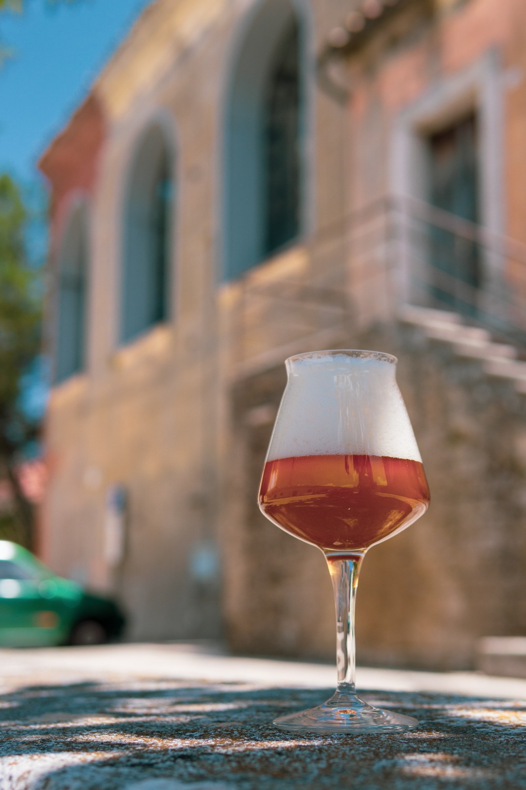 Calice di birra ambrata con schiuma bianca su un tavolo all'aperto con edificio sfocato sullo sfondo.
