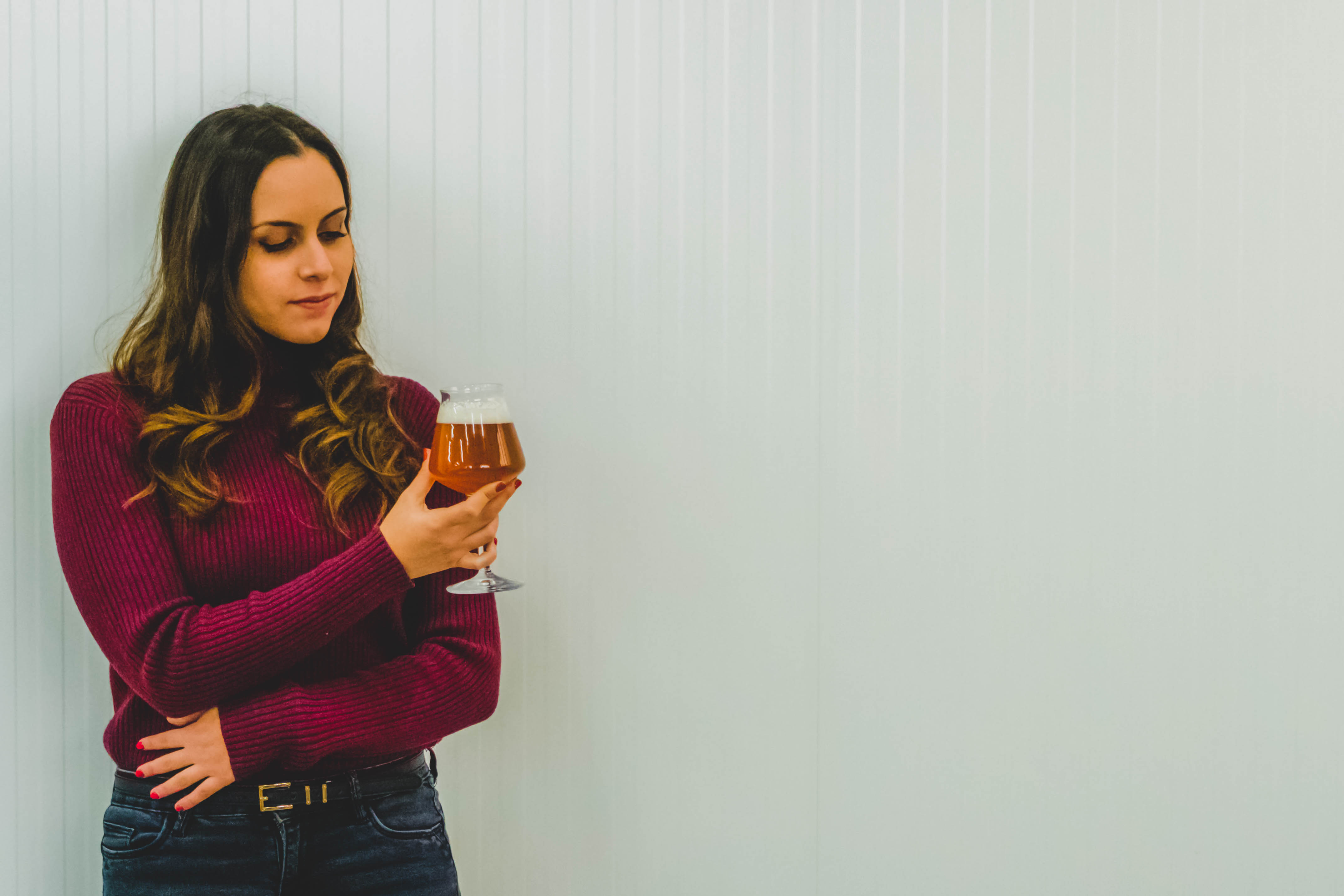 Donna con maglione bordeaux che osserva un bicchiere di birra chiara con schiuma.
