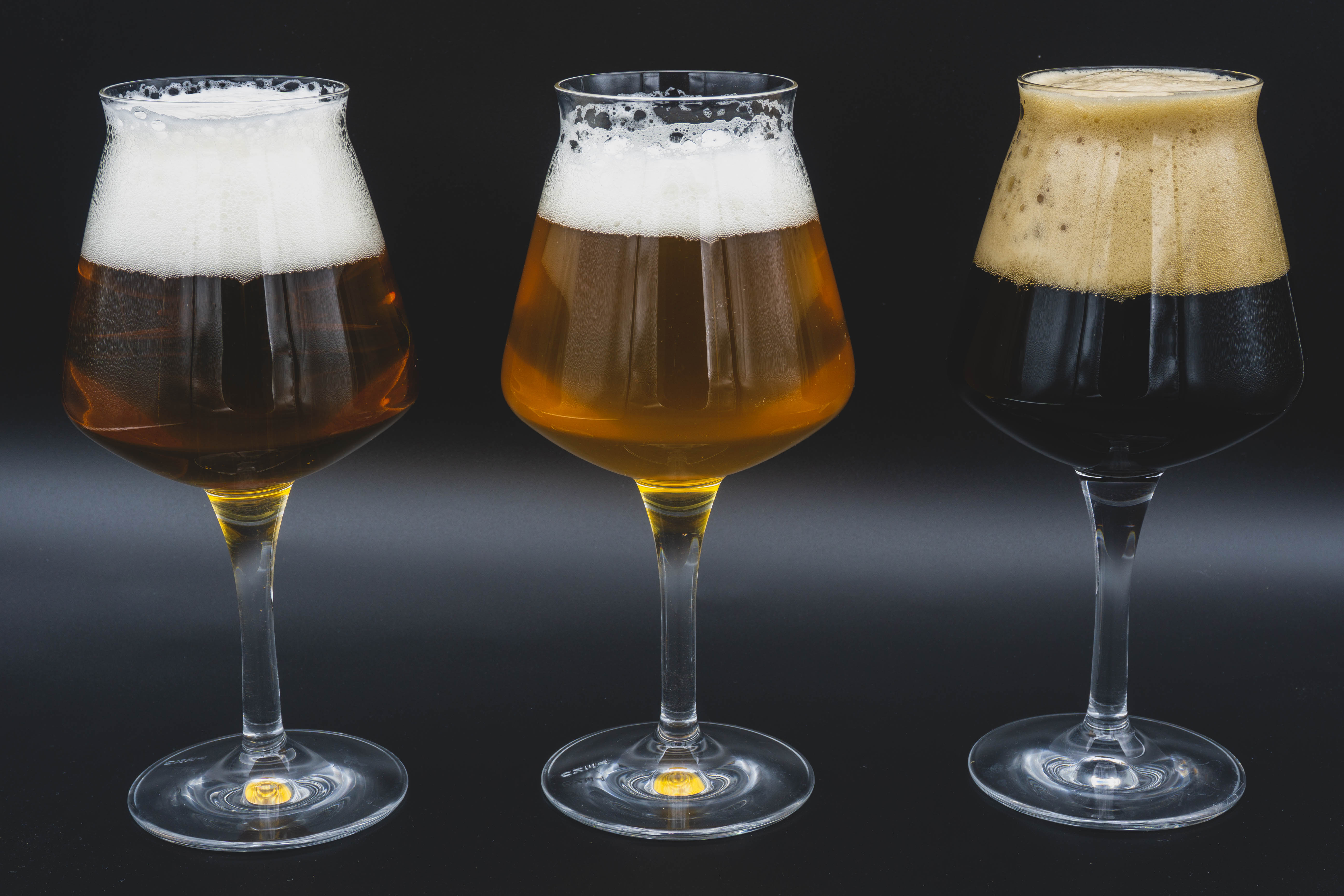 Tre calici di birra con schiuma, due di colore chiaro e uno scuro, su sfondo nero.