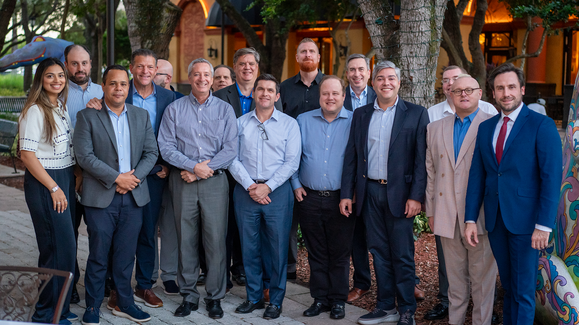 Group of fourteen people, mostly men in business attire, standing outdoors on a paved walkway with trees and an illuminated building in the background.