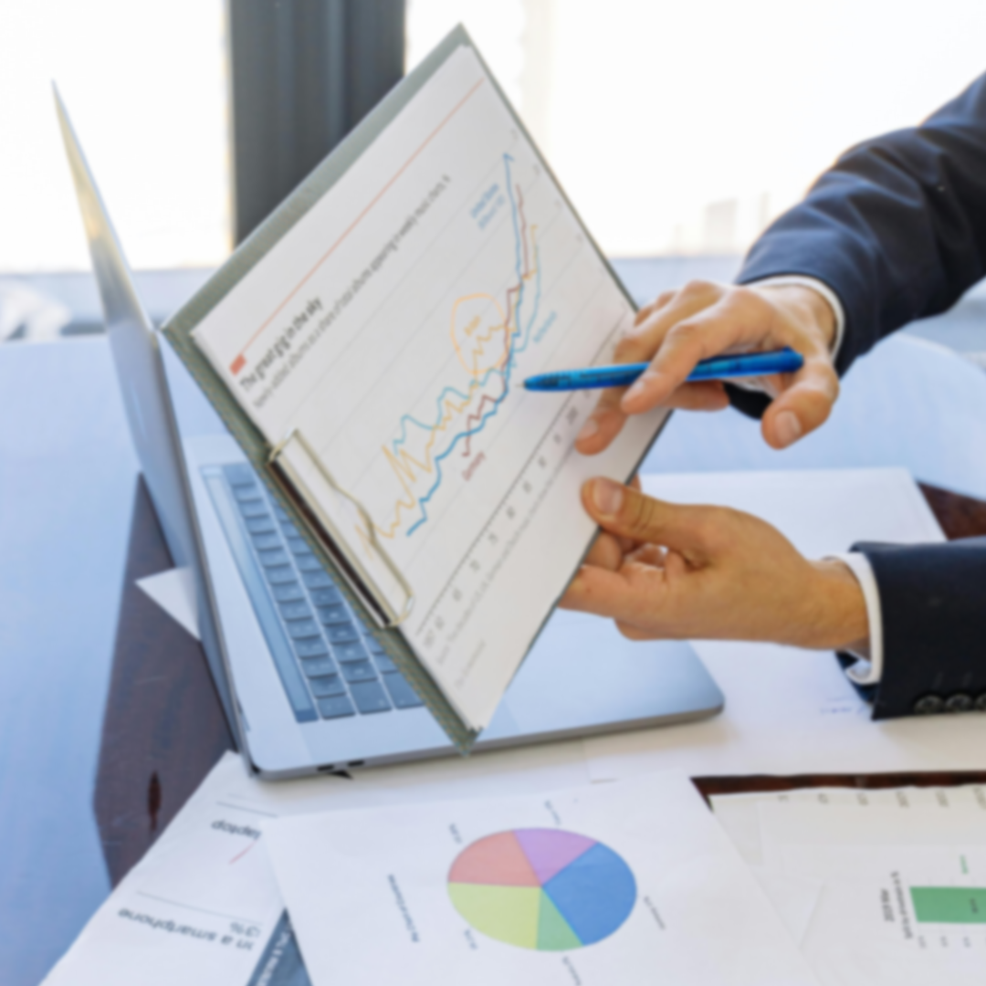 Person in business attire pointing with a pen to a line chart on a clipboard, with a laptop and printed pie and bar charts on a desk.