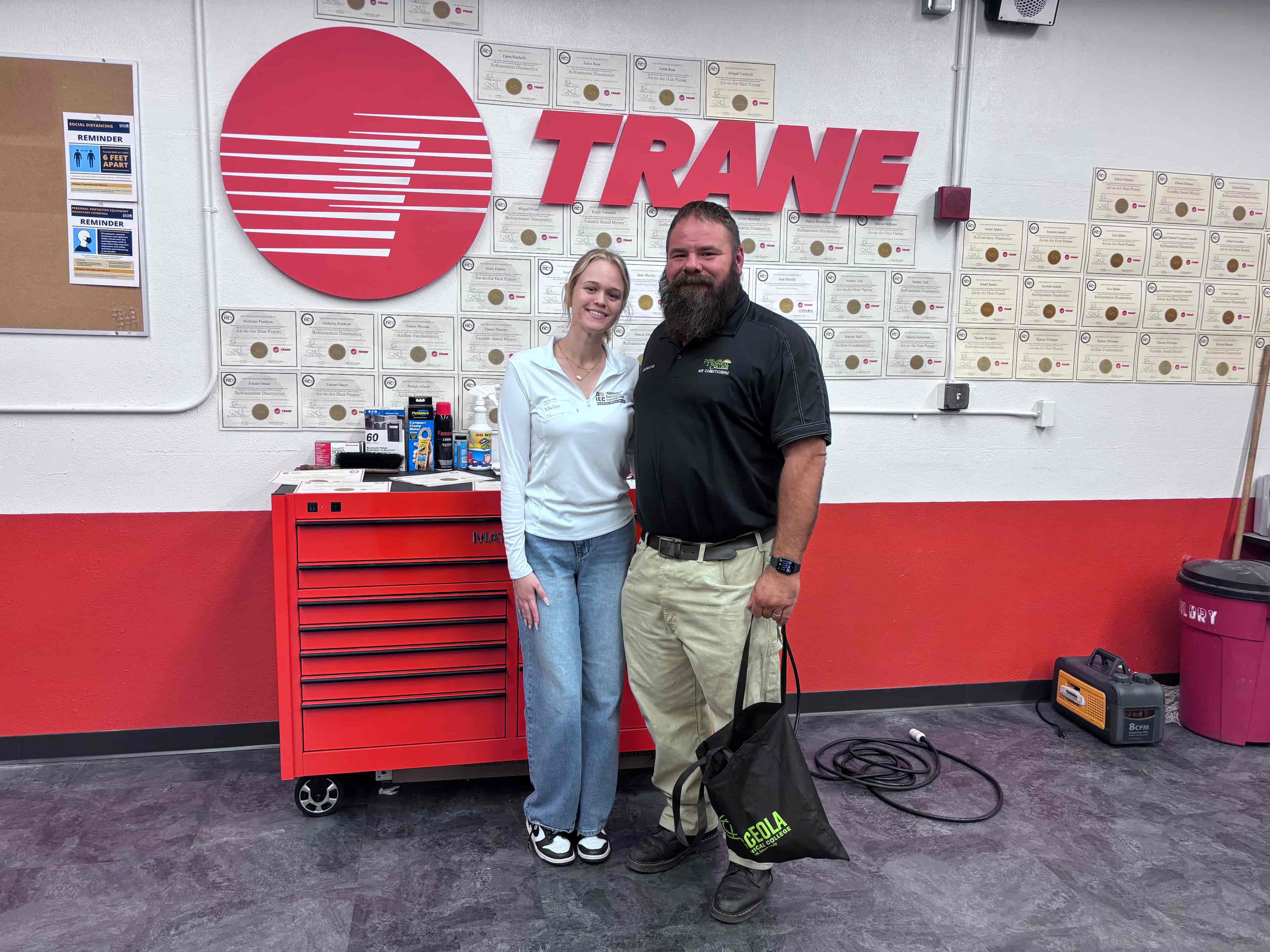 Isabella posing with instructor in front of wall of certificates and Trane logo