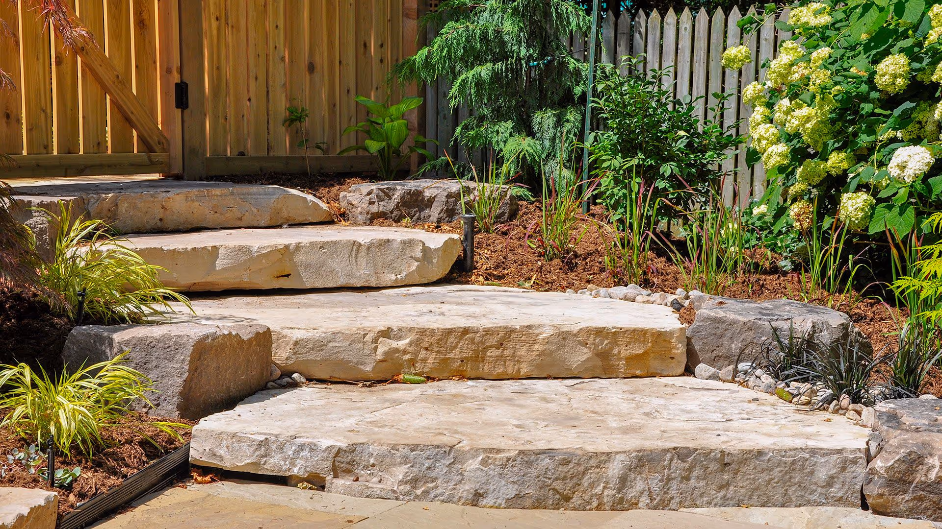 Stone steps near wooden fence surrounded by mulch and landscaping by Antonio's Outdoors