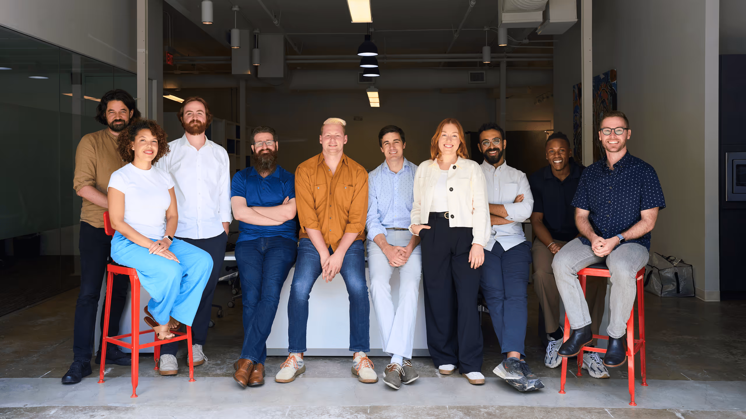 Diverse group of nine smiling professionals posing in modern office space, some seated on red stools and others standing.