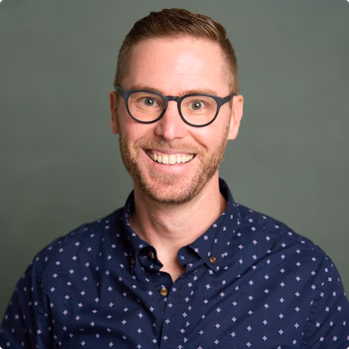 Photo of Trey Closson. Smiling man with short hair and glasses wearing a dark blue patterned button-up shirt against a green background.