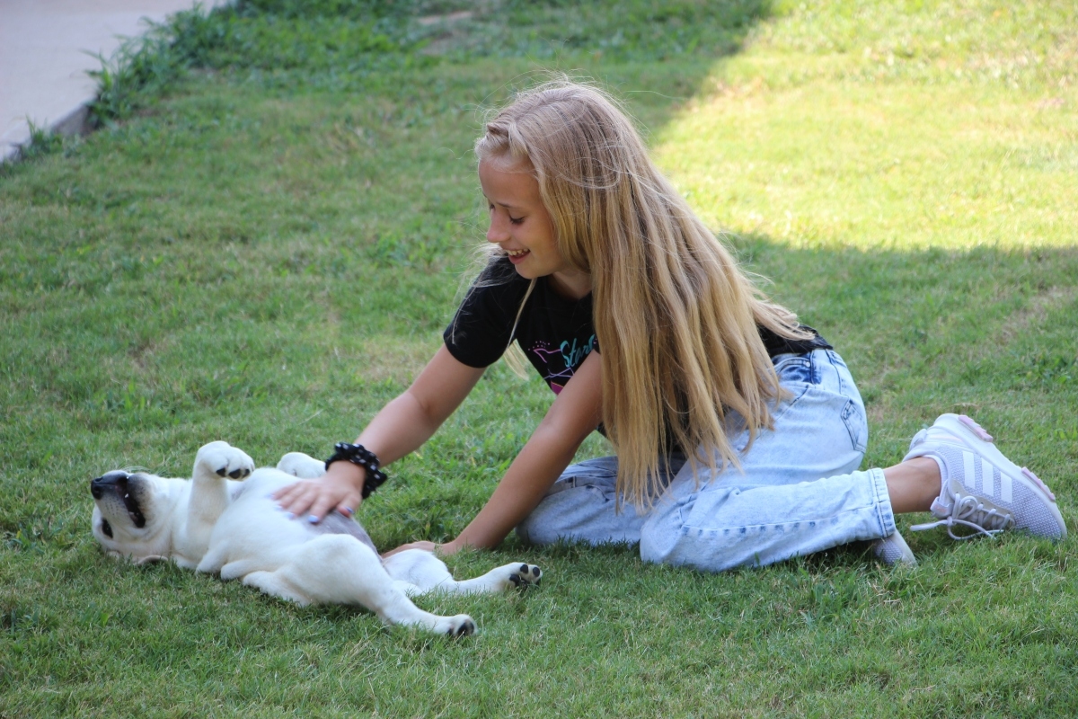 blonde girl sitting in grass rubbing a white puppies belly