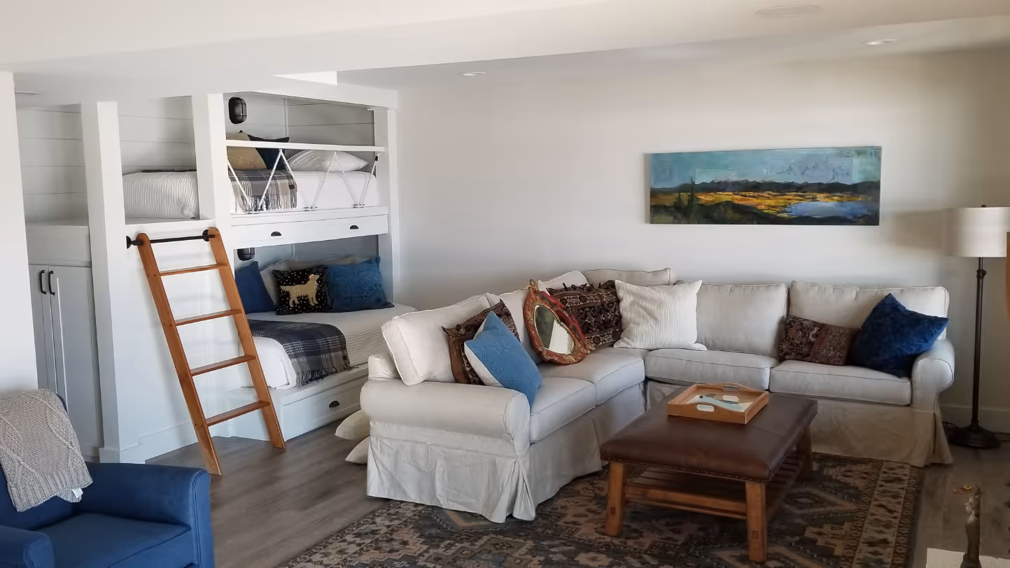Cozy living room with a white L-shaped sofa adorned with assorted pillows, a wooden coffee table, built-in bunk beds with a wooden ladder, a blue armchair, and a landscape painting on the wall.