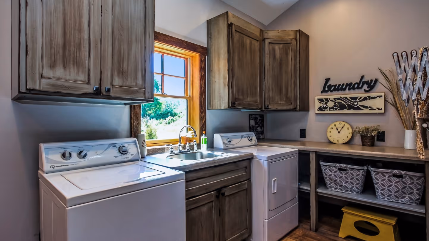 Laundry room with a washing machine, dryer, wooden cabinets, a window with natural light, a sink, decorative baskets, and a clock on the countertop.