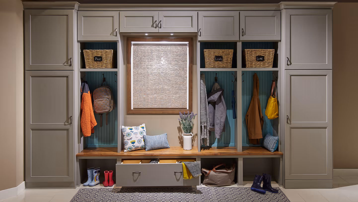 Organized mudroom with gray cabinets, wooden bench, labeled wicker baskets, coats, boots, and a window with a woven shade.