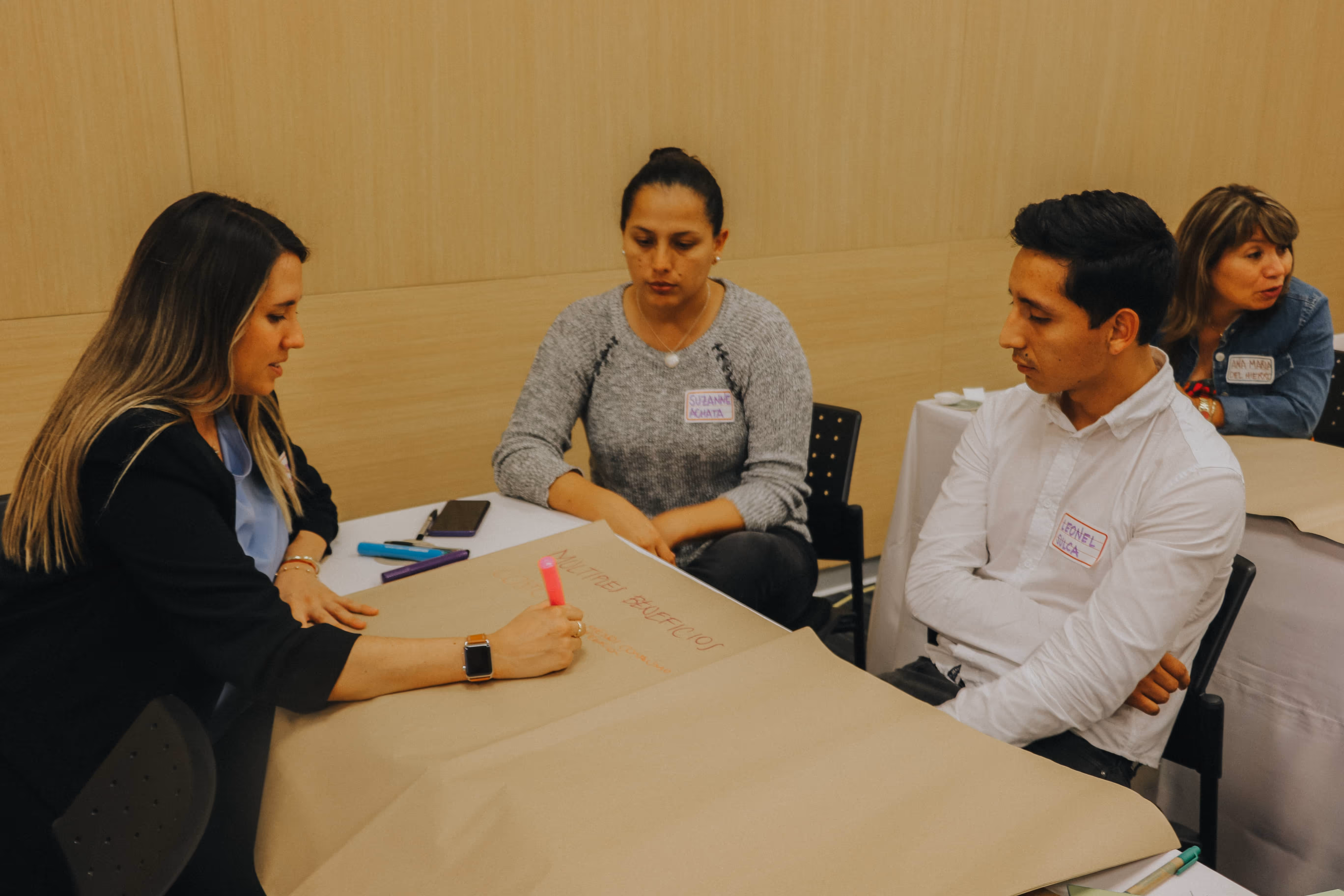 Cuatro personas sentadas en mesas, una mujer escribe con marcador rosa en un papel grande mientras los demás observan.
