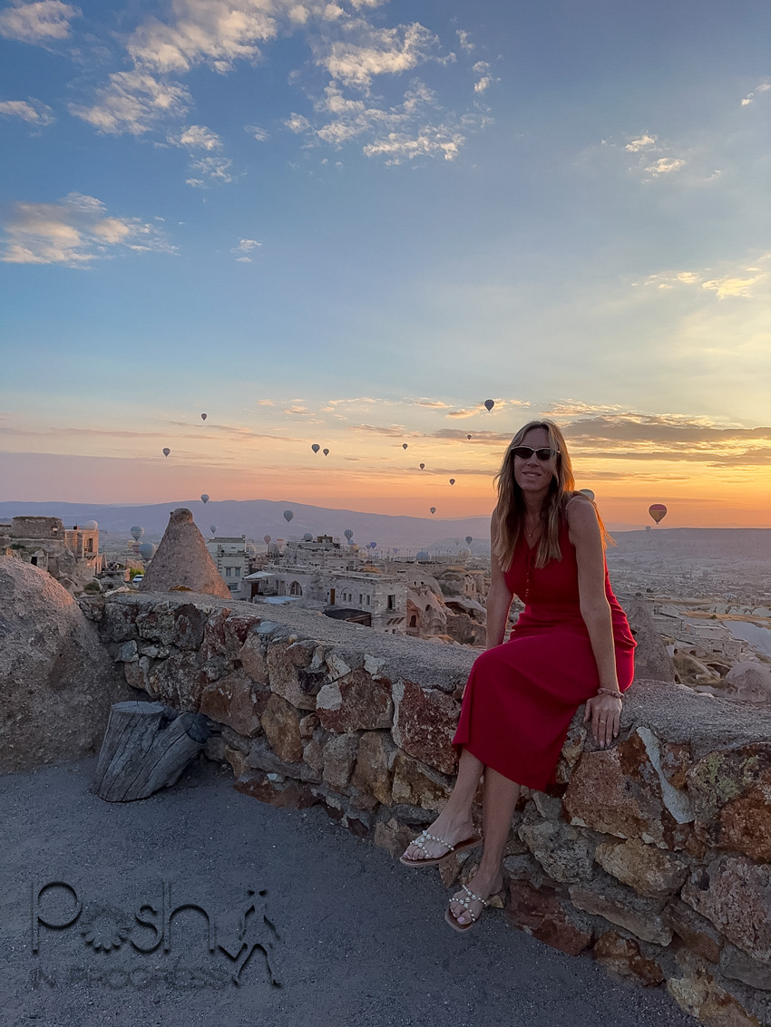 Woman in a red dress sitting on a stone wall at sunrise, with hot air balloons floating over a village in the distance