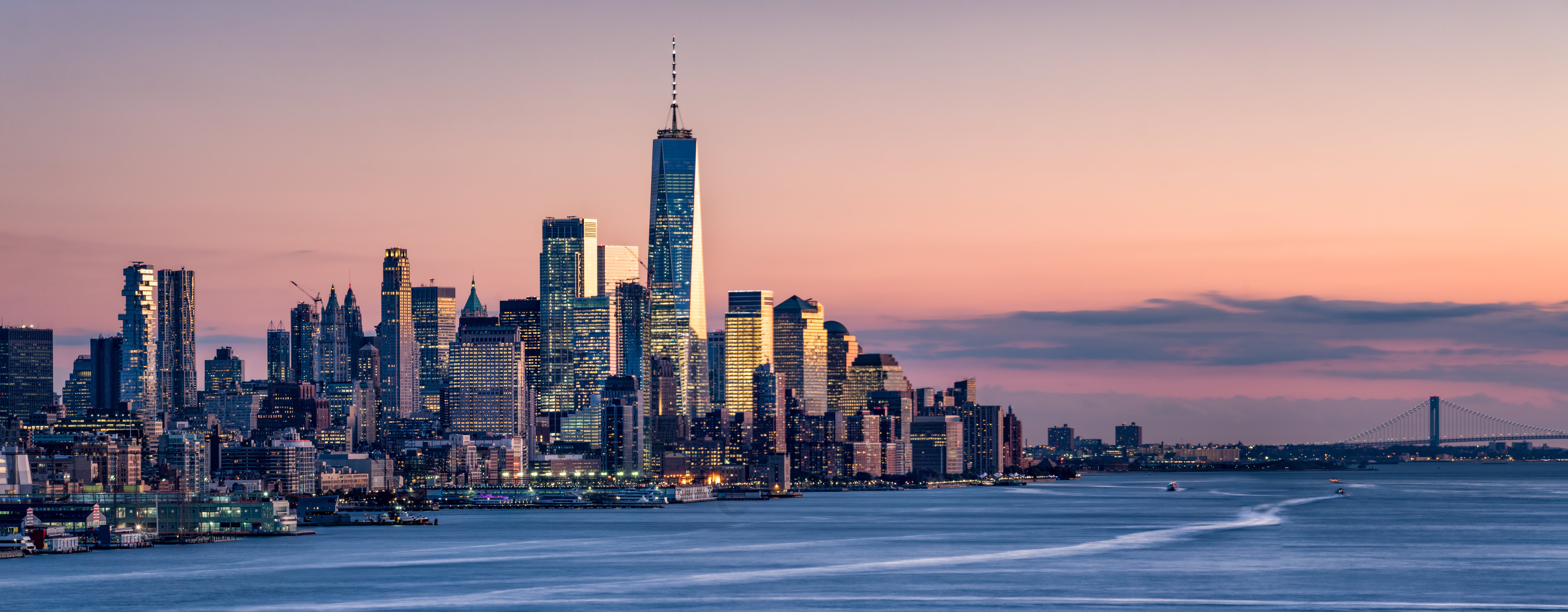 A photograph of the Lower Manhattan (New York City) skyline at sunset, with the One World Trade Center featured prominently. The sky is colored in orange and purple hues.
