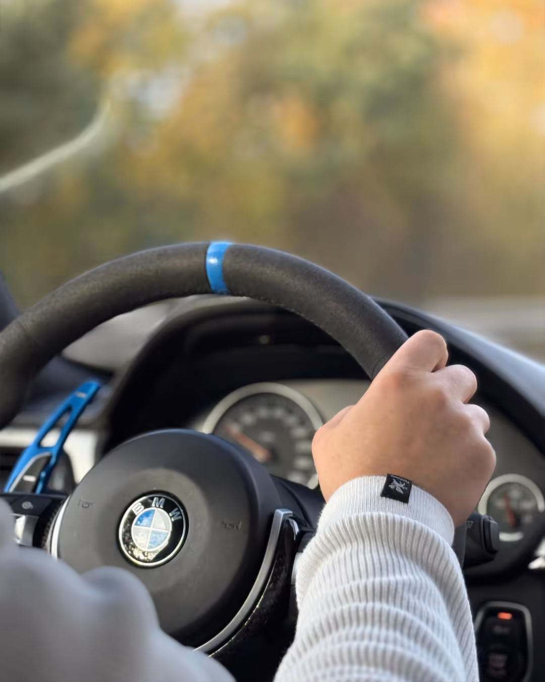 Hand in weißem Pullover am Lenkrad eines BMW mit blauem Akzent, unscharfer Hintergrund.