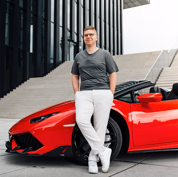 Mann in grauem T-Shirt und weißer Hose lehnt vor einem roten Sportwagen vor einem modernen Gebäude mit großen Treppen.