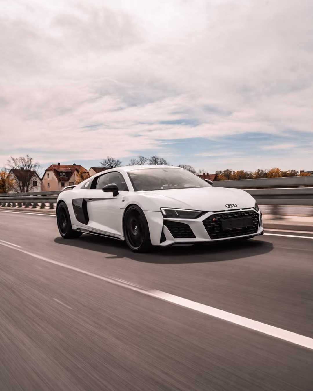 Weißer Audi Sportwagen fährt auf einer Straße mit Häusern und bewölktem Himmel im Hintergrund.