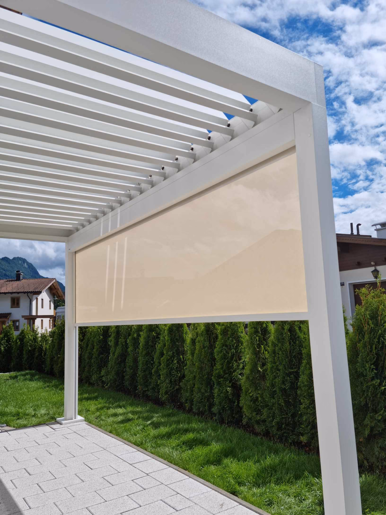 Weiße Pergola mit beigem Sichtschutz vor einer grünen Hecke und einem sonnigen Garten.