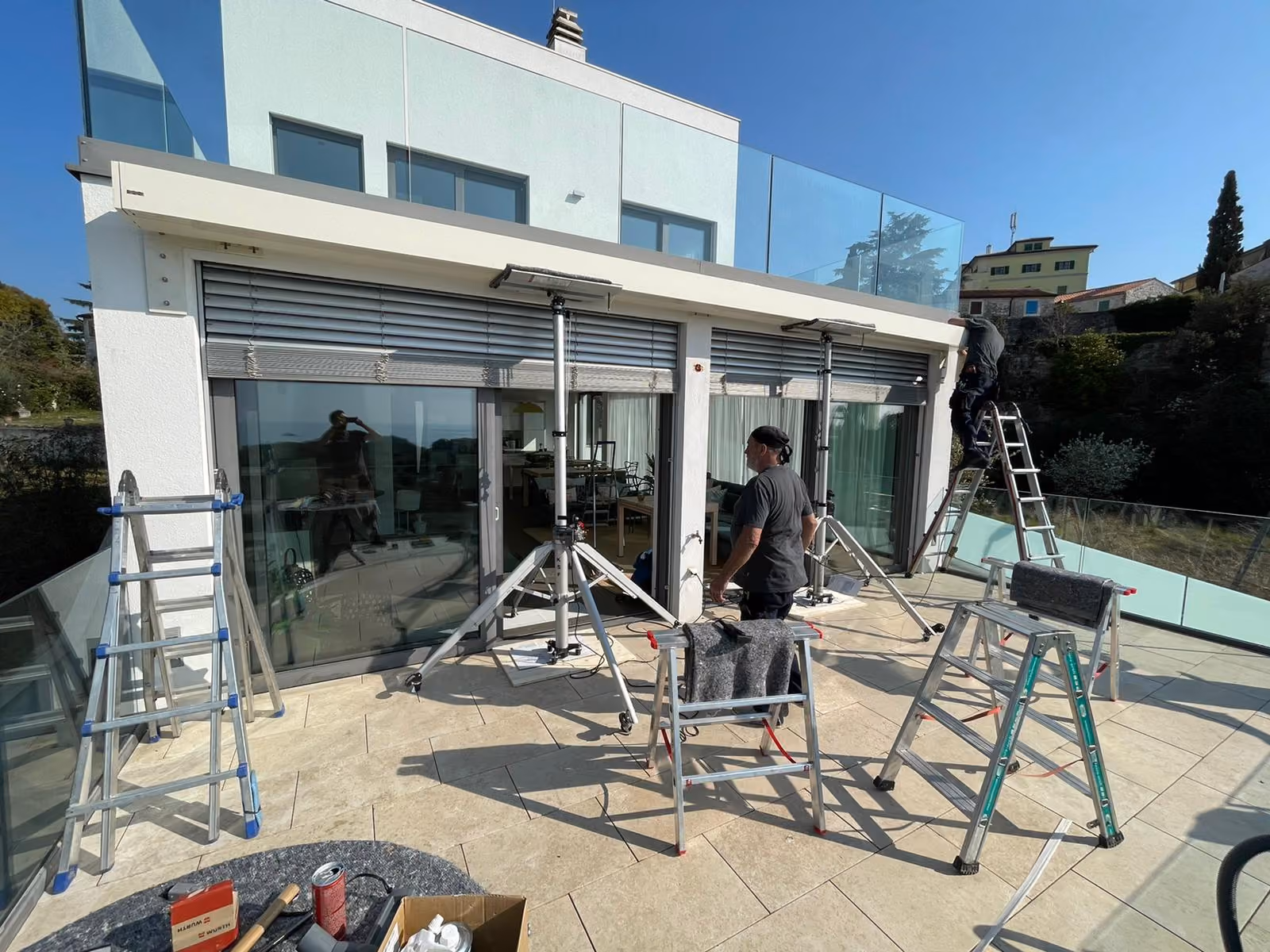Zwei Arbeiter auf einer Terrasse montieren Rollläden an einem modernen weißen Haus bei Sonnenschein.