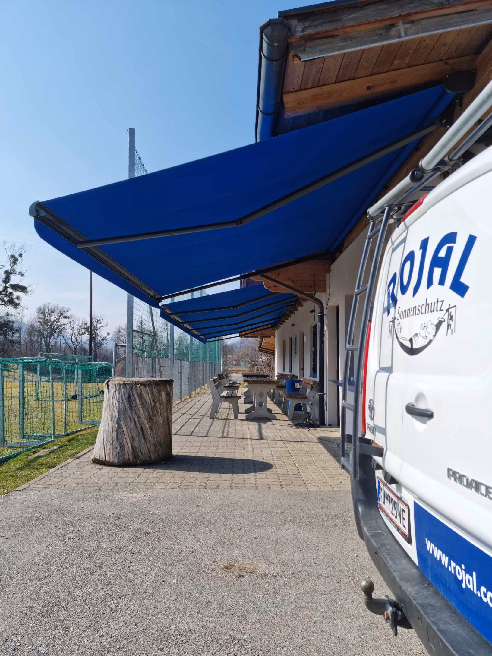 Terrasse mit blauen Markisen, Bänken und einem Stapel Holz neben einem weißen Van mit Rojal Werbung.