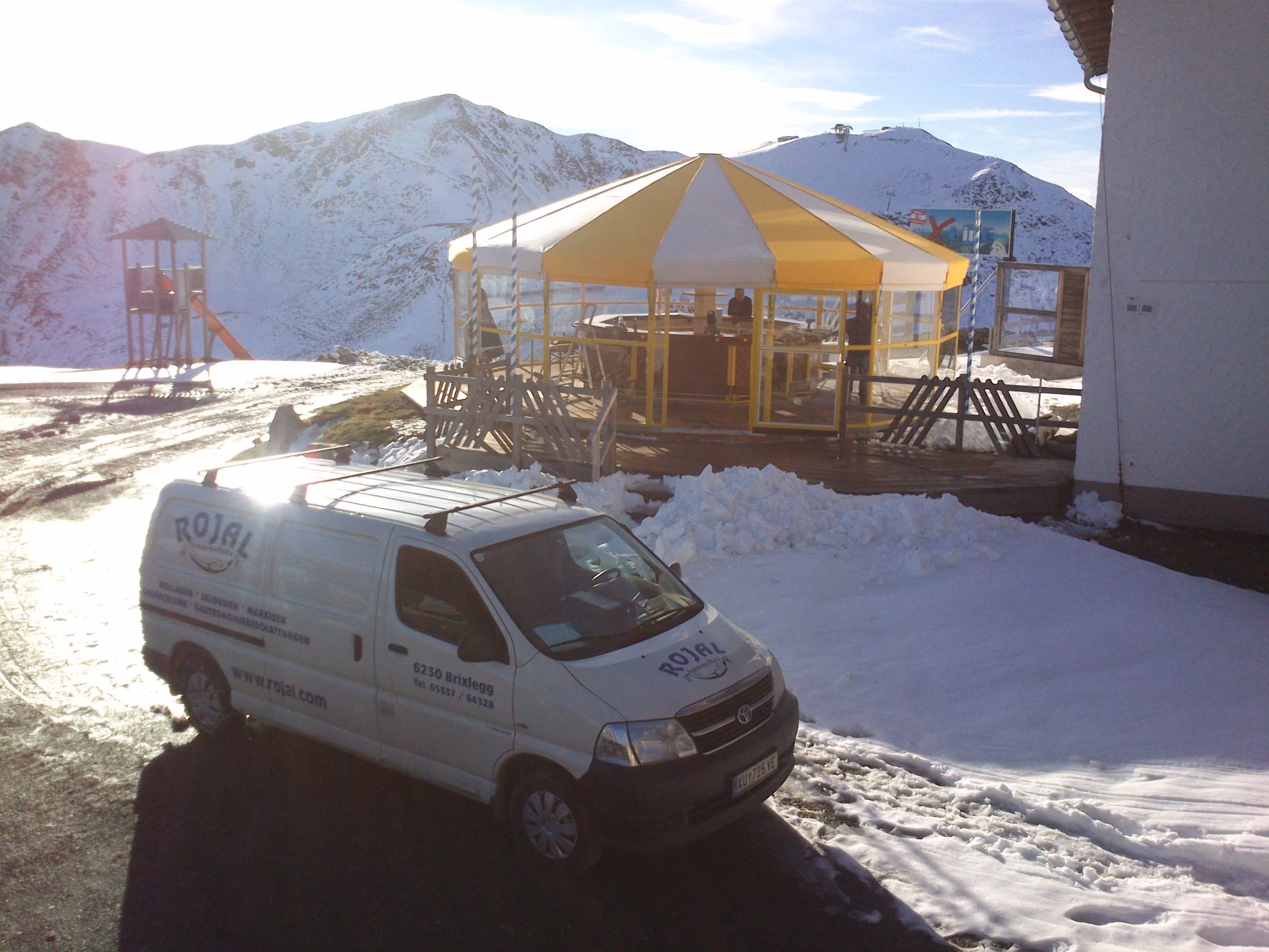 Weißer Lieferwagen vor gelb-weißem Zelt im Schnee mit schneebedeckten Bergen im Hintergrund.
