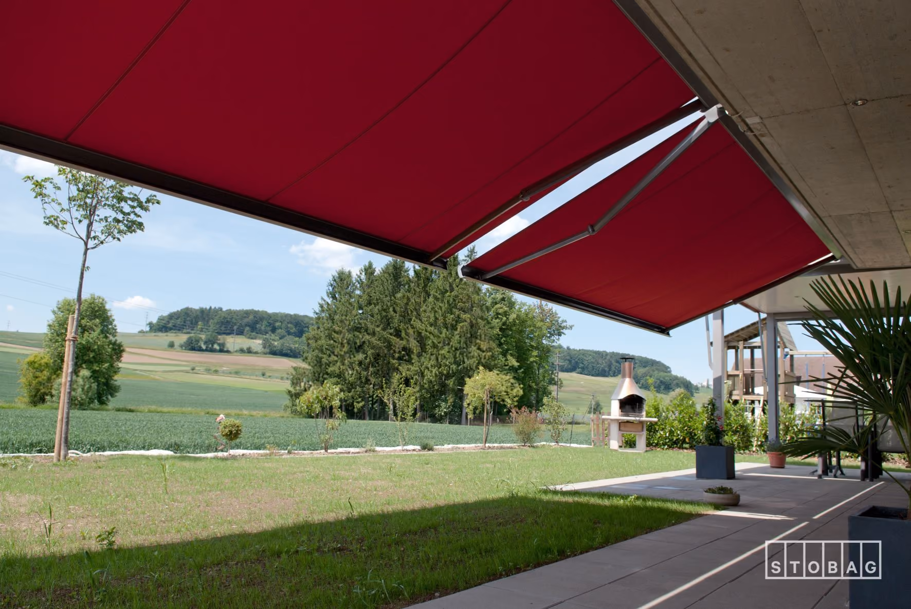 Rote Markisen spenden Schatten auf einer Terrasse mit Blick auf grüne Felder und Wald.