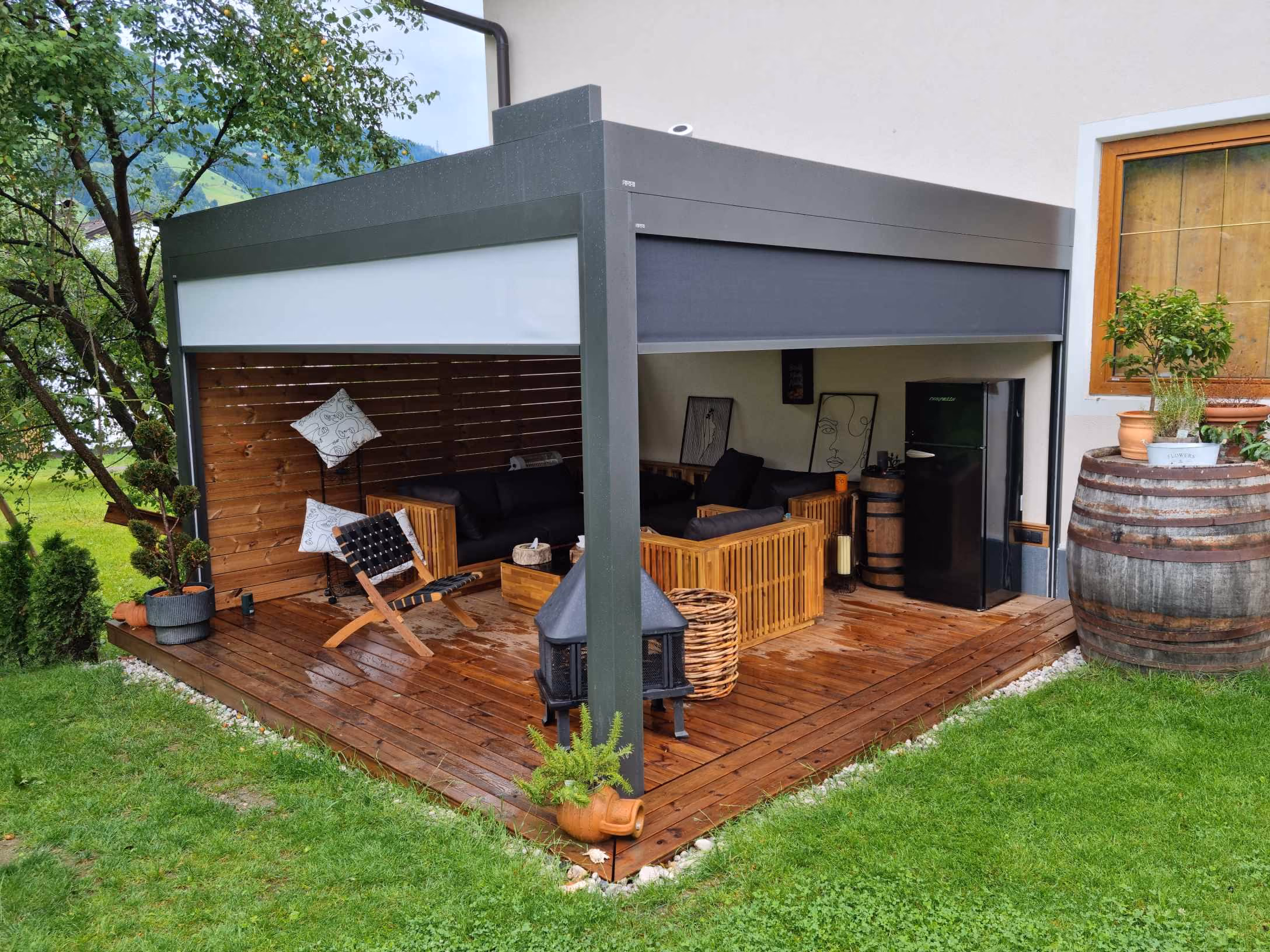 Überdachte Holzterrasse mit Lounge-Möbeln, Kaminofen und Kühlschrank, umgeben von grünem Gras.