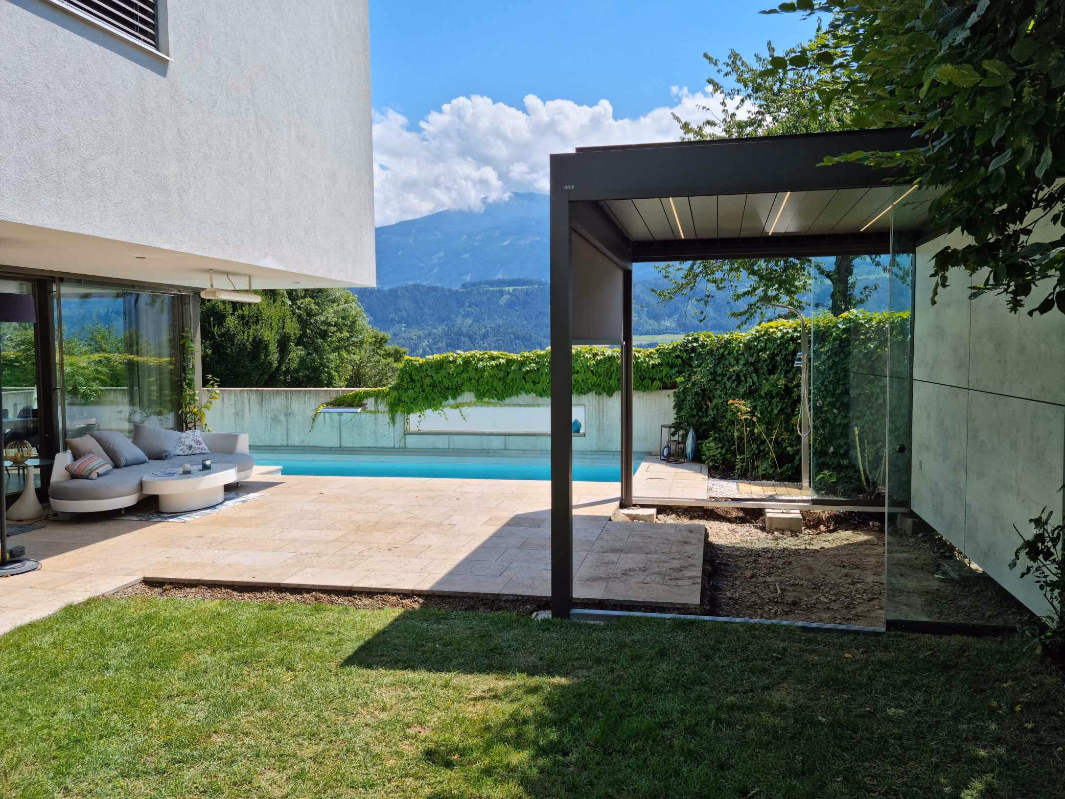 Moderner Garten mit Rasen, überdachtem Außenbereich, Pool und Bergblick bei Sonnenschein.