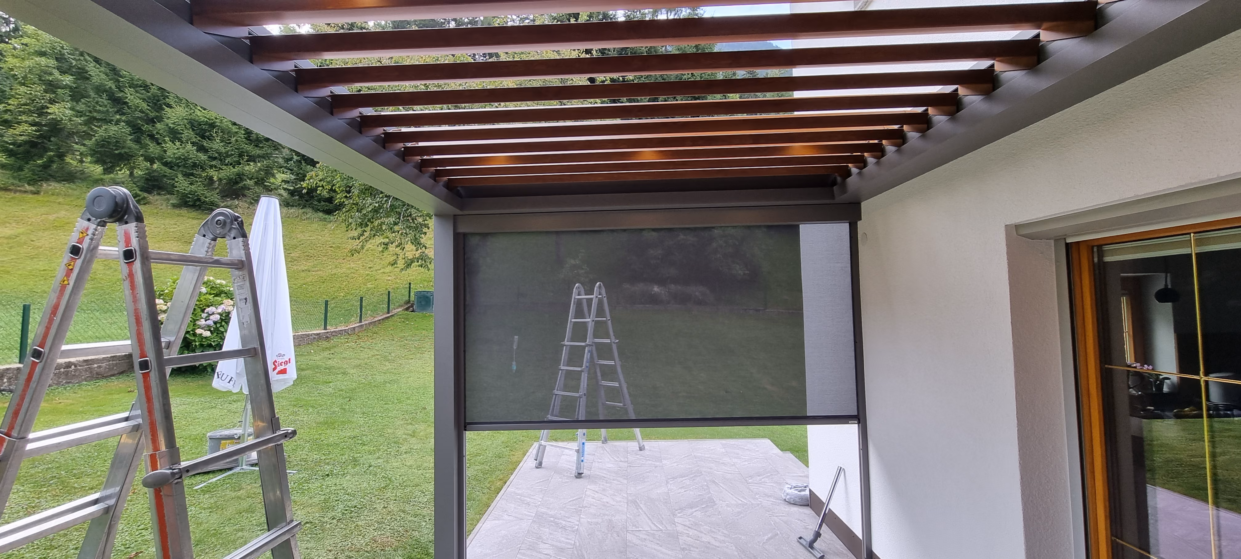 Terrassenüberdachung mit Holzlamellen und Leiter neben einem grauen Sonnenschutzrollo.