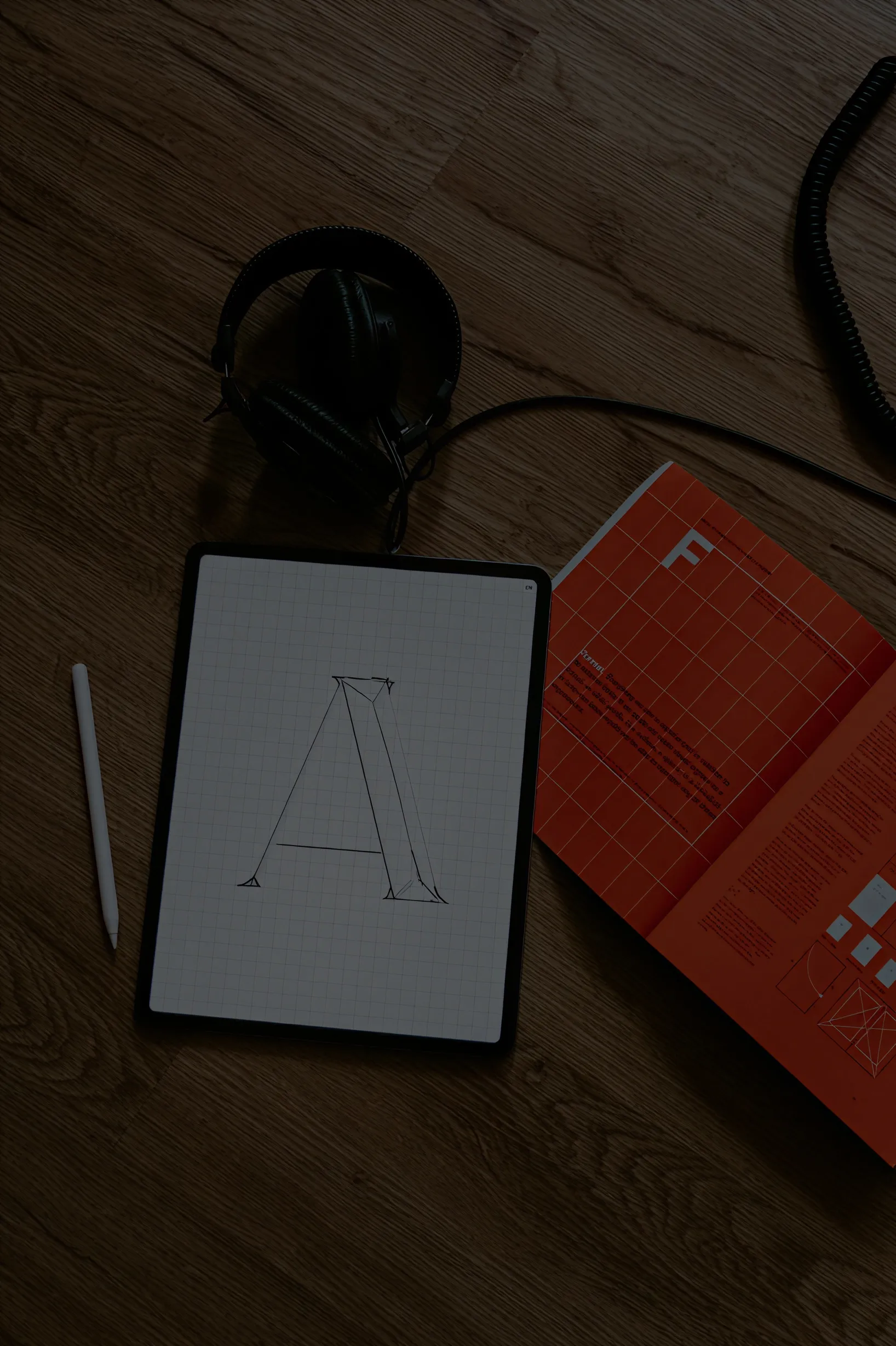 Tablet on wooden floor displaying a grid with a sketch of a capital letter A, surrounded by a white stylus, black headphones, and an open red book.