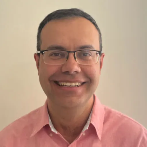 Smiling man with glasses and short dark hair wearing a pink collared shirt against a plain background.