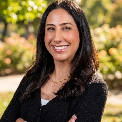 Smiling woman with long dark hair wearing a black sweater, standing outdoors with blurred greenery behind her.