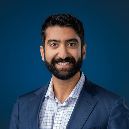 Smiling man with dark hair, beard, wearing a blue suit jacket and checkered shirt, blue background.