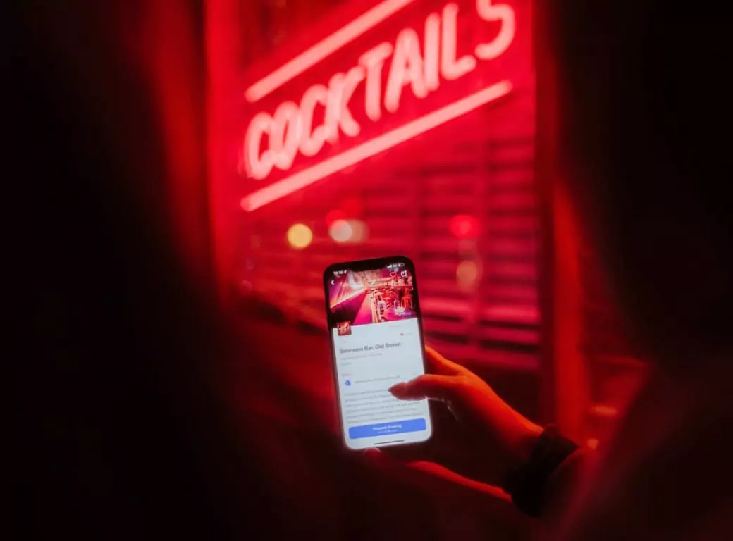 A user holding their phone in front of a neon “Cocktails” sign, exploring Ambl’s curated list of the best nightlife spots nearby.