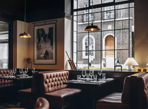 A stylish and intimate restaurant with leather booth seating, elegant table settings, and large windows overlooking a classic brick building—one of the premium dining spots available for instant booking through the Ambl app.