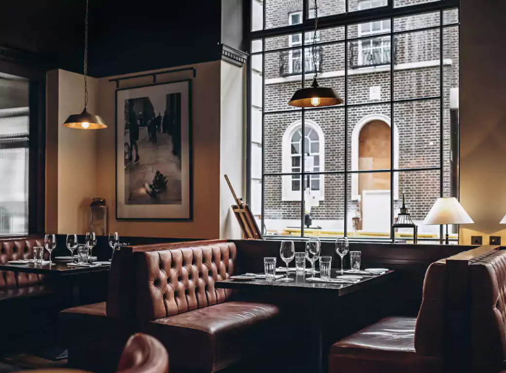 A stylish and intimate restaurant with leather booth seating, elegant table settings, and large windows overlooking a classic brick building—one of the premium dining spots available for instant booking through the Ambl app.