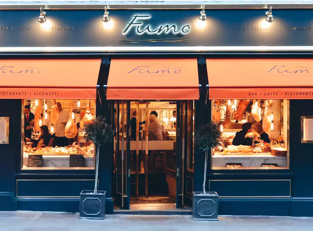The elegant exterior of Fumo, an Italian restaurant and bar with warm lighting, inviting décor, and orange awnings, offering a vibrant dining experience. Available for instant booking through the Ambl app.
