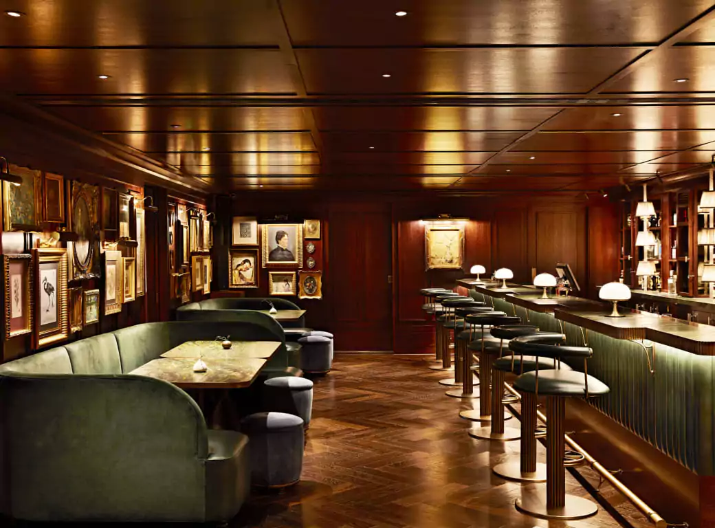 A sophisticated, dimly lit bar with plush green seating, a long elegant counter, and framed artwork adorning the walls. One of the exclusive venues available through the Ambl app.