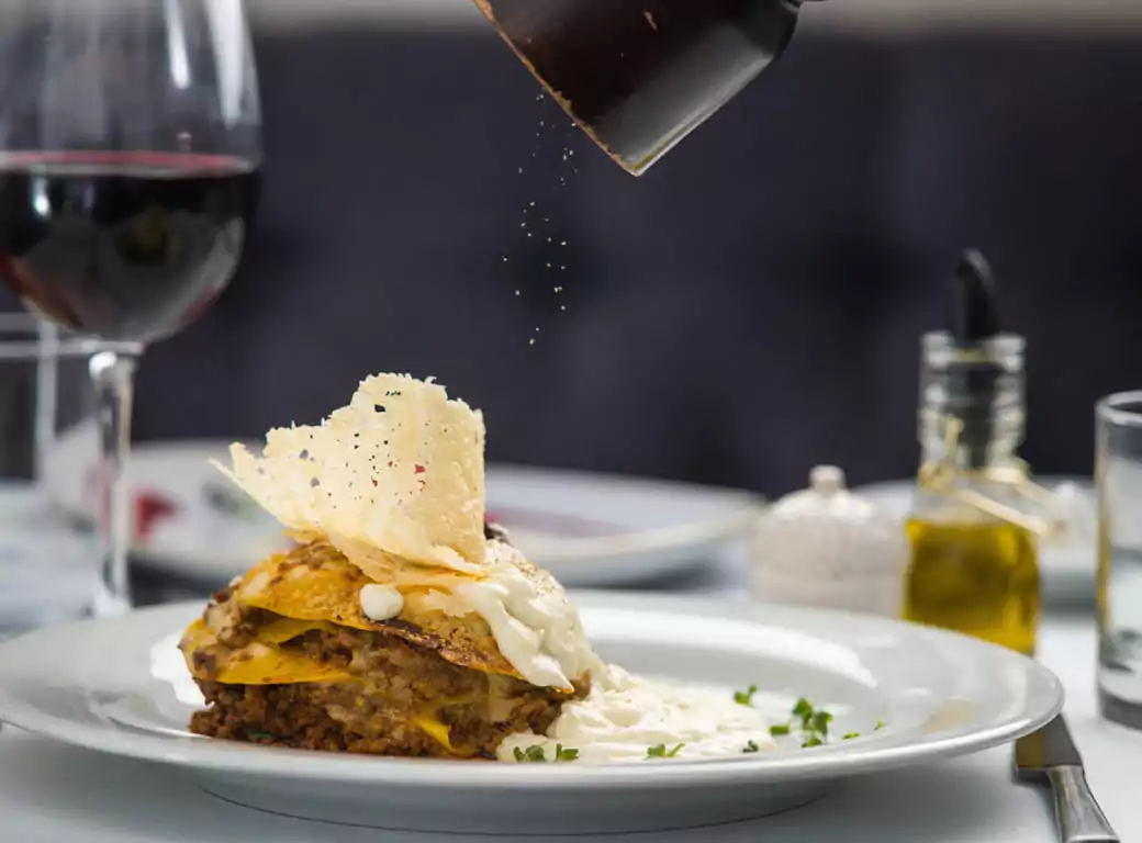A gourmet lasagna dish being freshly seasoned with black pepper, served with a glass of red wine at an upscale Chelsea restaurant. Discover the best dining spots in Chelsea with Ambl for last-minute reservations.