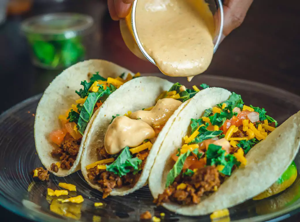 A plate of three soft tacos filled with seasoned meat, fresh greens, diced tomatoes, and shredded cheese, topped with a creamy sauce being drizzled from above. Discover the best spots for mouth-watering Mexican cuisine with Ambl.