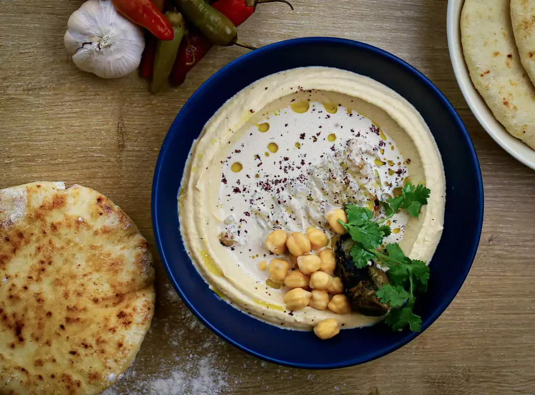 A bowl of creamy hummus garnished with whole chickpeas, roasted eggplant, fresh parsley, and a drizzle of olive oil, served alongside warm pita bread and pickled peppers. Explore the best Mediterranean dining experiences with Ambl.
